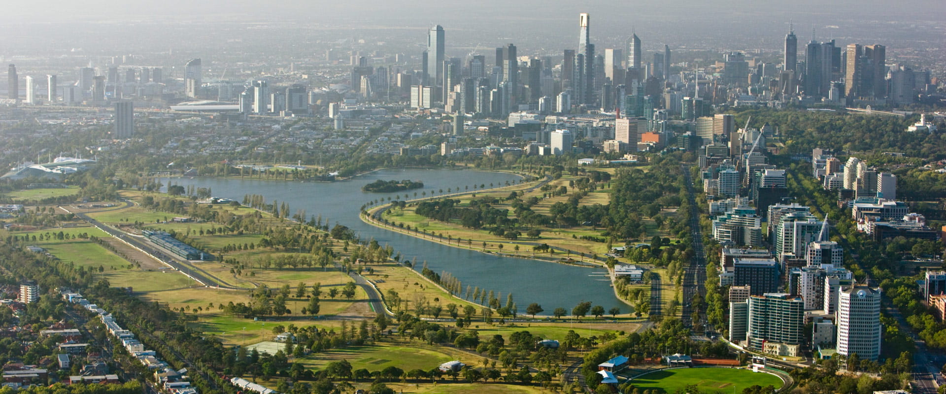 Aerial view of Albert Park lake lined by trees with the Melbourne city skyline in the background.